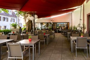 Außengastronomie mit, grauen Stühlen und Tischen, die unter einem roten Sonnensegel stehen. Auf der linken Seite ragt ein mächtiger Baumstamm empor, im Hintergrund befindet sich die Glasfront des Restaurants.
