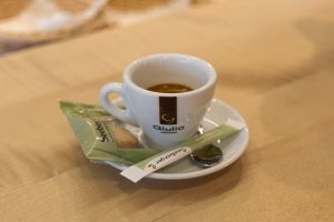 Eine weiße Espressotasse der Marke „Giulio“ mit Kaffee auf einer Untertasse. Daneben liegen ein verpackter Seeberger-Keks und ein Zuckerbriefchen.