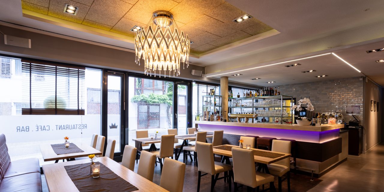 Der Sitzbereich des Restaurants an einer großen Fensterfront. Braune Lederbänke und beige Stühle stehen an Holztischen. Auf der Scheibe ist spiegelverkehrt „RESTAURANT . CAFÉ . BAR“ zu lesen.