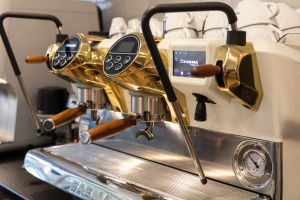 Nahaufnahme einer eleganten, weiß-goldenen Faema-Siebträgermaschine mit Holzgriffen. Auf der Oberseite sind weiße Kaffeetassen zum Vorwärmen gestapelt.