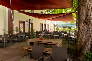 Restaurantterrasse mit Tischen und Stühlen. Darüber zwei große, Sonnensegel, die an der Hauswand befestigt sind. Ein dicker Baumstamm flankiert die rechte Seite.
