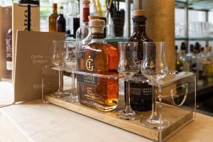 Eine Flasche „Giare Amarone“ Grappa und eine weitere dunkle Flasche stehen zusammen mit vier passenden Grappagläsern auf einem Holzaufsteller mit Acrylhalterung auf der Theke.