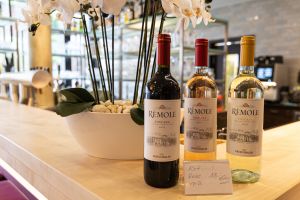 Drei Weinflaschen der Marke „Rémole Toscana“ (Rot, Rosé und Weiß) stehen nebeneinander auf einer Holztheke vor einer weißen Orchidee. Ein kleines Schild zeigt den Preis von 18 Euro.