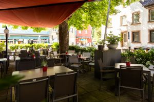 Perspektive auf den Randbereich der Terrasse. Mehrere Tische und Stühle stehen im Schatten eines großen Baumes und eines Sonnensegels. Auf den Tischen sind Pflanzentöpfe mit Tischnummern platziert. Im Hintergrund sind Häuser sichtbar.
