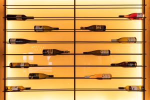 Eine modern gestaltete Weinwand mit horizontalen Metallstreben vor einem warmen, gelb beleuchteten Hintergrund. Mehrere Weinflaschen liegen sortiert auf den Halterungen.