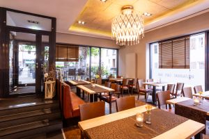 Blick vom Restaurantbereich in Richtung der verglasten Eingangstür. Holzstühle und -bänke stehen an fertig gedeckten Tischen unter einem auffälligen, geometrischen Kronleuchter.